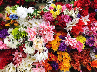 A vibrant assortment of artificial flowers in a variety of colours and styles, including lilies, roses, daisies, and mixed foliage, displayed on a shop wall.
