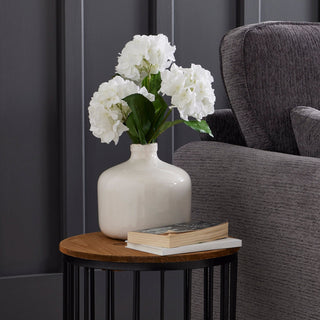 White ceramic vase with faux hydrangeas on a round wood and metal side table next to a grey armchair.