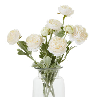 Bunch of artificial ranunculus stems with pale apricot flowers and green foliage, shown in a simple glass vase.
