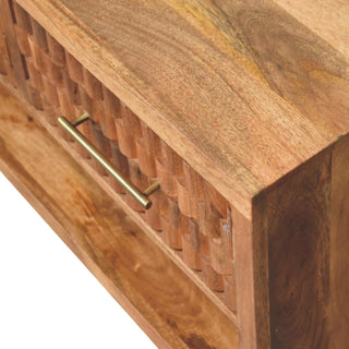 High angle detail of the golden teak top surface, carved drawer front, brass handle, and open shelf.