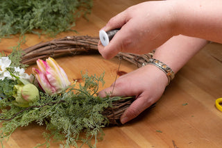 A person's hands attaching the artificial asparagus fern foliage to a wicker wreath base using fine wire.