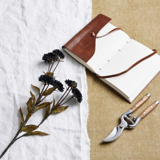 Artificial black chrysanthemum stem on white linen, beside an open leather journal and metal gardening shears.