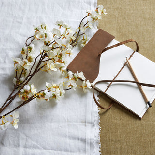 Artificial white Japanese blossom branch styled on white linen next to a leather-bound notebook and pencil on a brown surface.