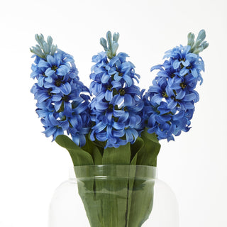Three blue artificial hyacinth stems grouped in a clear glass vase against a white backdrop.