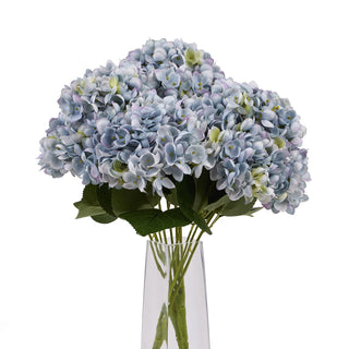 Artificial blue hydrangea bunch filling a tall, clear glass vase, isolated on a white studio background.