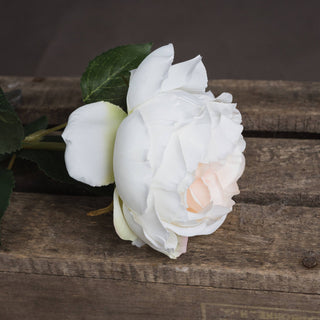 Close-up of the artificial rose bloom showing white and pale blush petals resting on dark wood.