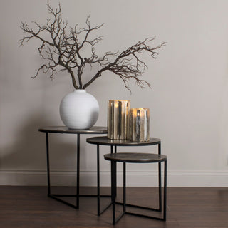 Dark artificial branches arranged in a round white vase on a set of three metal nesting tables.
