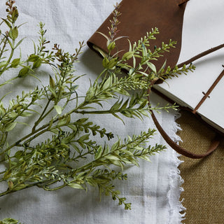 Close-up detail of the artificial broom stem showing variegated green leaves and buds, next to a brown notebook.