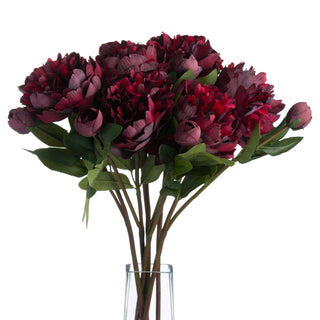 A bunch of artificial burgundy peony roses and buds with green leaves, stems in a clear vase.