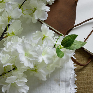 Close-up of white artificial cherry blossoms and green leaves against a brown leather notebook and fringed white linen.