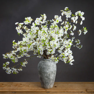 Faux white cherry blossom sprays arranged in a textured grey vase, set on a wooden table against a dark grey background.