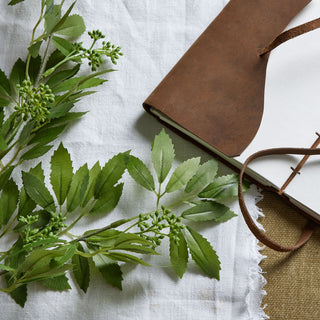 Close-up of the artificial fennel stem's serrated leaves and green berries, next to an open leather notebook.