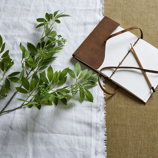 Artificial fennel stem on white linen next to a brown leather notebook and pencil, overhead shot.