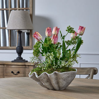 Artificial gypsophila mixed with faux tulips and foliage in a large, ribbed shell planter on a wooden table.