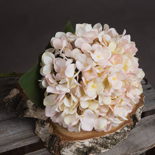 Artificial hydrangea bloom resting on a round birch slice, showing textured cream and blush petals.