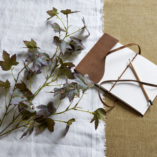 Artificial variegated maple leaf stem with green and purple foliage, styled with a notebook on linen.