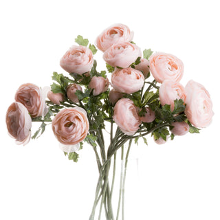 Artificial pastel pink ranunculus stems and buds arranged in a clear glass vase on a white background.