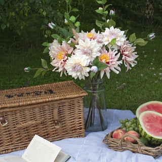 Artificial dahlias arranged in a vase on a picnic blanket next to a wicker hamper, fruit, and an open book.
