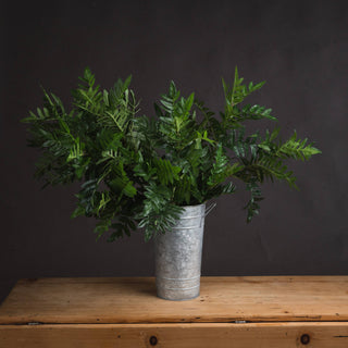 Artificial green foliage bunch in a metal bucket on a wooden table, shot front-on against a dark grey background.