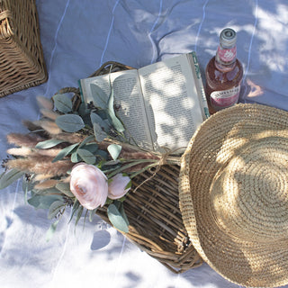 Artificial ranunculus in a mixed bouquet on a wicker tray with a book and straw hat at a picnic.