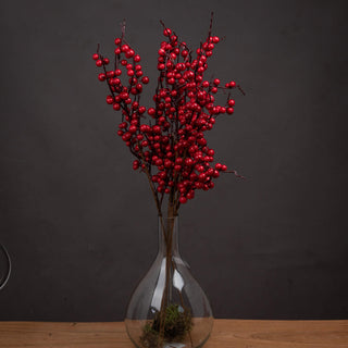 Artificial red berry picks displayed in a glass vase on a wooden surface against a dark grey background.
