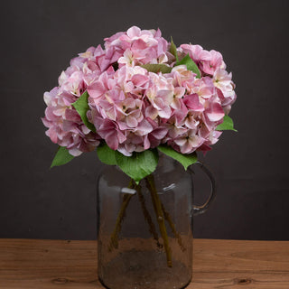 Artificial pink hydrangea head with green leaves in a textured grey glass jug on a wooden surface.