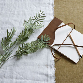 Artificial silver wattle stem lying on white fabric next to a brown leather journal.