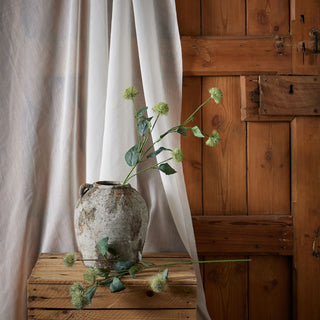Artificial thistle stem in a rustic urn on a wooden crate, with sheer curtains and a wooden door backdrop.