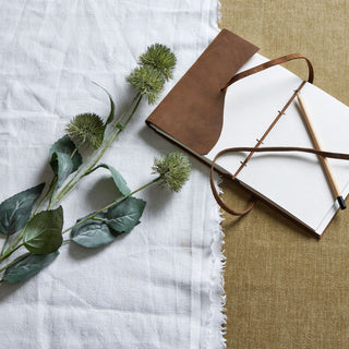 Artificial thistle stem on white linen next to an open brown notebook and pencil on a khaki surface.