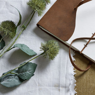 Detail shot of two artificial thistle stems lying on white fabric beside an open brown leather notebook.
