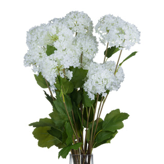 Artificial white viburnum bouquet with green foliage in a clear glass vase against a white background.