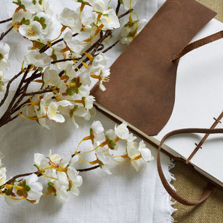 Textured close-up showing white artificial blossom petals against brown stems, white linen, and brown leather notebook.