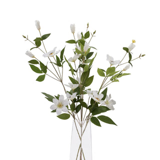 Multiple artificial white clematis stems with green foliage arranged in a tall, clear glass vase.