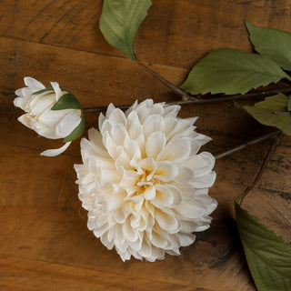 Detailed view of the cream dahlia bloom and bud, showing fabric petals and green leaves against a wood grain background.