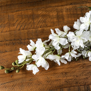 White artificial delphinium stem showing open flowers and green buds, laid diagonally on a rustic wooden surface.