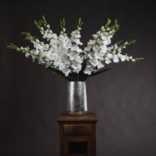 Full arrangement of white artificial gladioli in a silver vase, displayed on a small wooden cabinet.