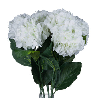 Group of artificial white hydrangeas and green leaves shown in a vase on a white background.