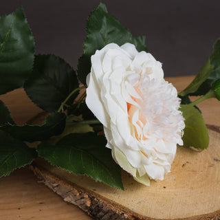 A close-up of the artificial white rose bloom and green leaves resting on a round wooden slice.