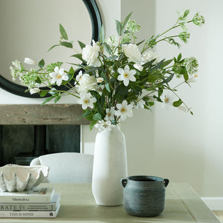 White roses and mixed artificial flowers arranged in a tall white vase on a dining table.