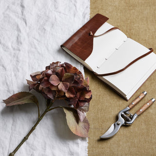 Artificial autumn brown hydrangea stem styled with a leather journal and secateurs on white and hessian fabric.