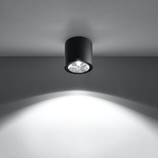 Black cylindrical ceiling light fixture illuminating a wall with a bright, focused downward beam.