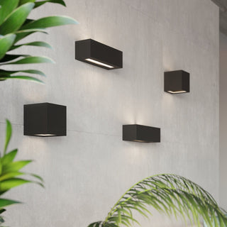Four black cubic and rectangular wall lights illuminating a grey tiled wall, framed by large green plants.