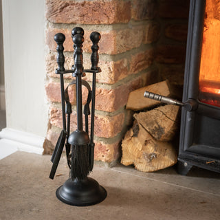 Black companion set next to a lit wood stove and stacked logs against a brick hearth.