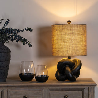 Styled shot of the lamp on a wooden sideboard, lit up, next to a vase of foliage and two glass tumblers.
