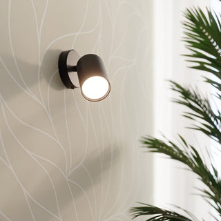 Black cylindrical wall lamp illuminated against light patterned wallpaper, with dark green houseplant leaves visible.