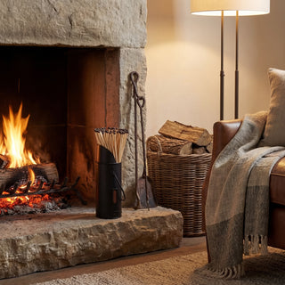 Black match holder on a stone hearth beside a lit fireplace, fire tools, and a wicker log basket.