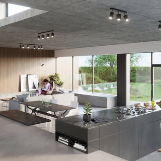 Black six-spot track lights installed on a concrete ceiling in a modern open-plan kitchen and living area.