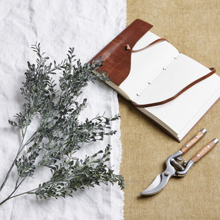 Blackened grass spray stem laid on white linen, next to an open leather notebook and a pair of wooden-handled secateurs.