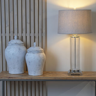 Two distressed white ginger jars displayed on a wooden console table next to a tall glass lamp.