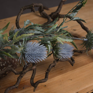 Close-up of two blue thistle heads and green foliage resting on wood, surrounded by dark, twisted branches.
